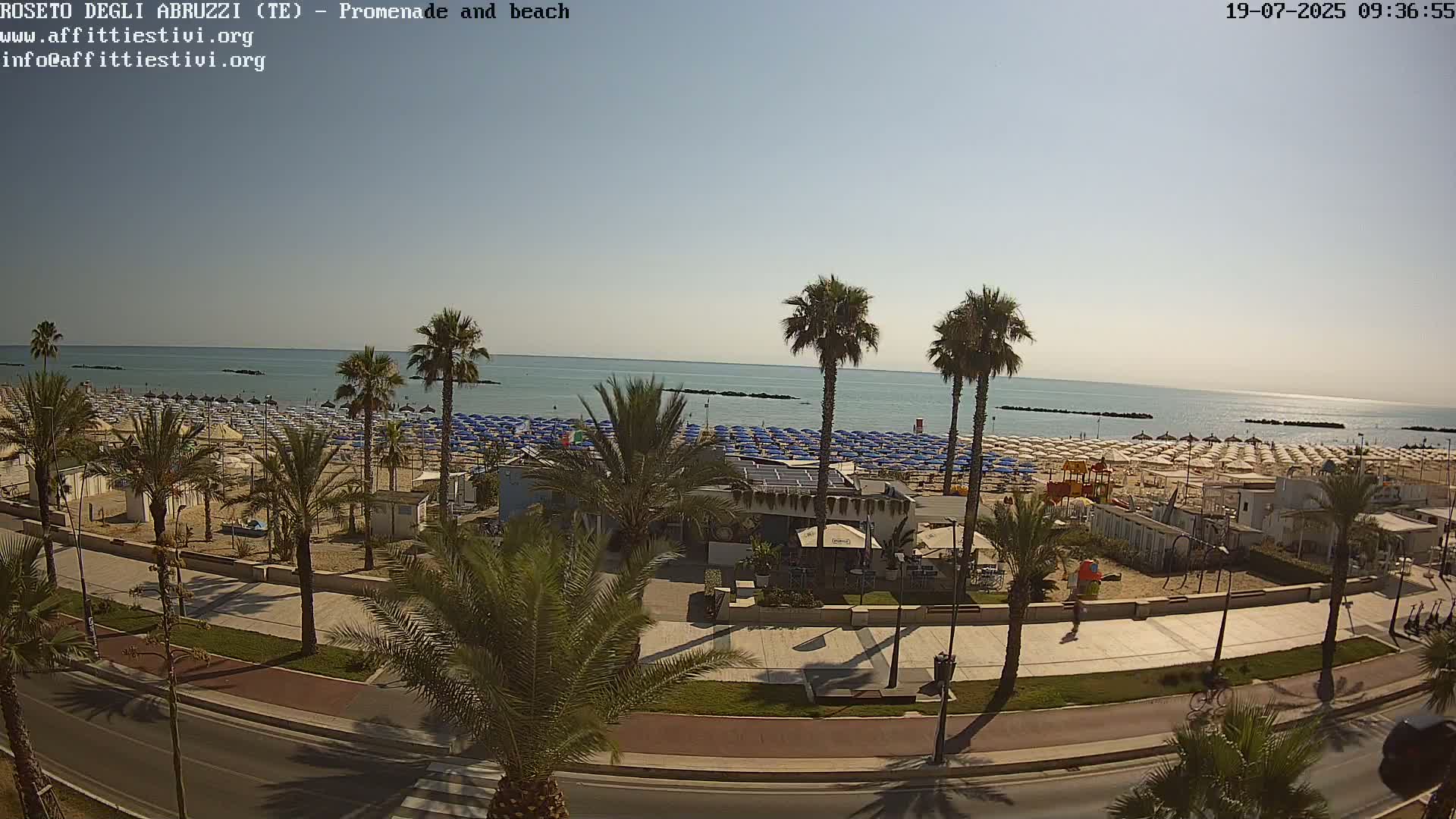 A sunny day at a beach, showing a promenade lined with palm trees, beach umbrellas, and a beachfront restaurant, overlooking a calm sea.