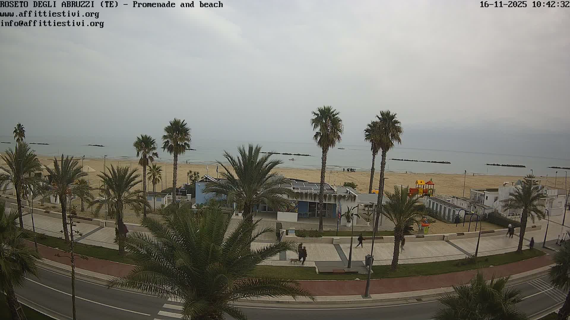On a cloudy day, a coastal promenade with palm trees and a road runs alongside a sandy beach featuring beach clubs, a playground, scattered people, and breakwaters in the calm sea.