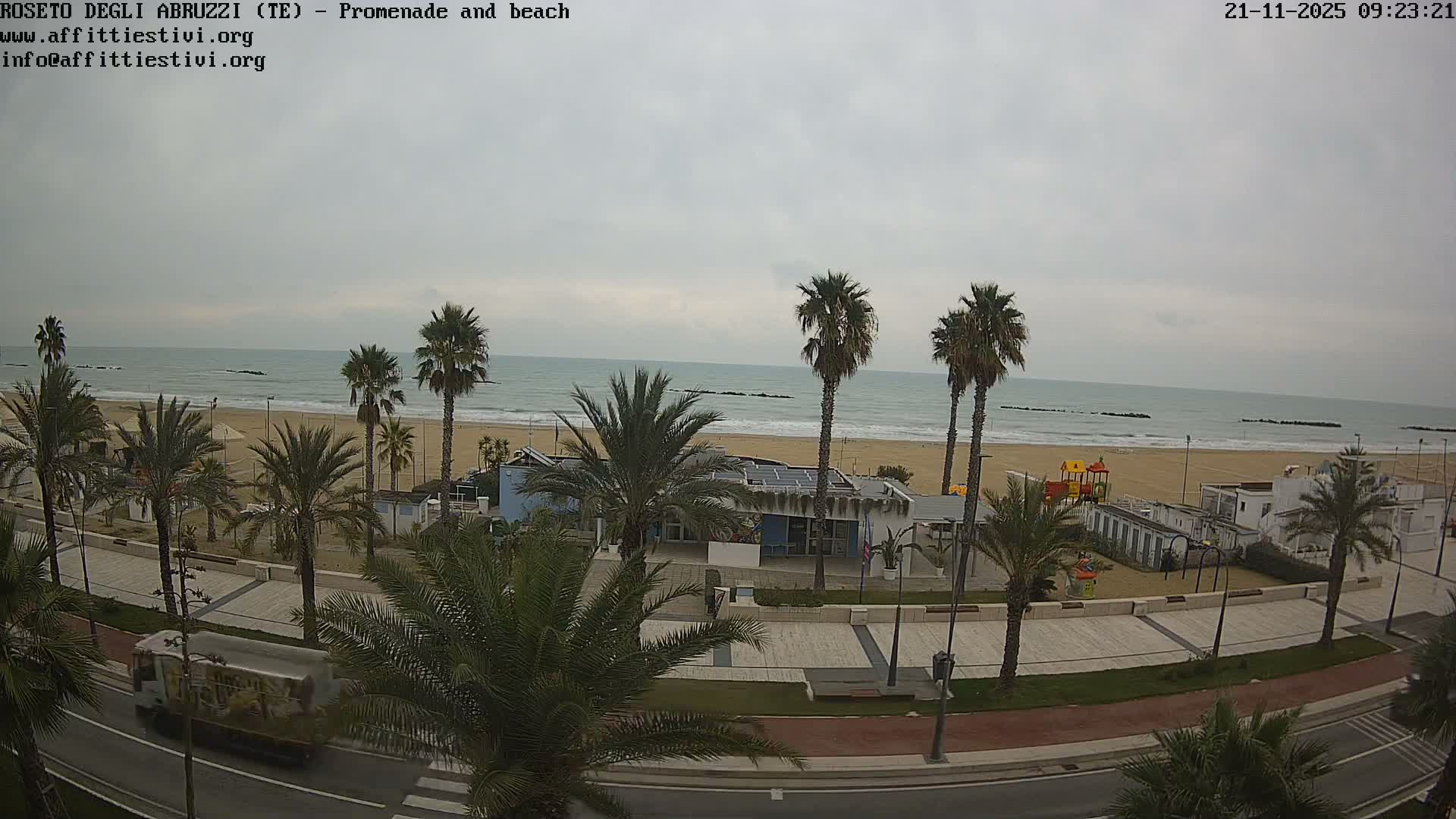 An overcast sky hangs over a coastal scene featuring a sandy beach with a playground, the calm sea with distant breakwaters, and a palm-tree-lined promenade alongside a road with a moving truck.