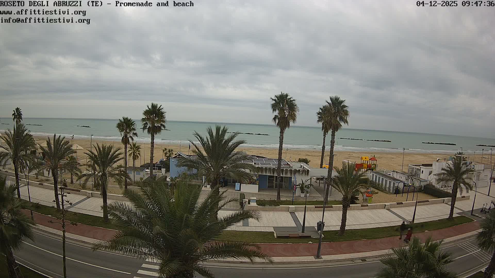 Under an overcast sky, a wide-angle view captures a coastal scene with a palm-lined promenade and road in the foreground, leading to a sandy beach dotted with buildings and a playground, all set against a calm, pale blue-green sea with distant breakwaters.