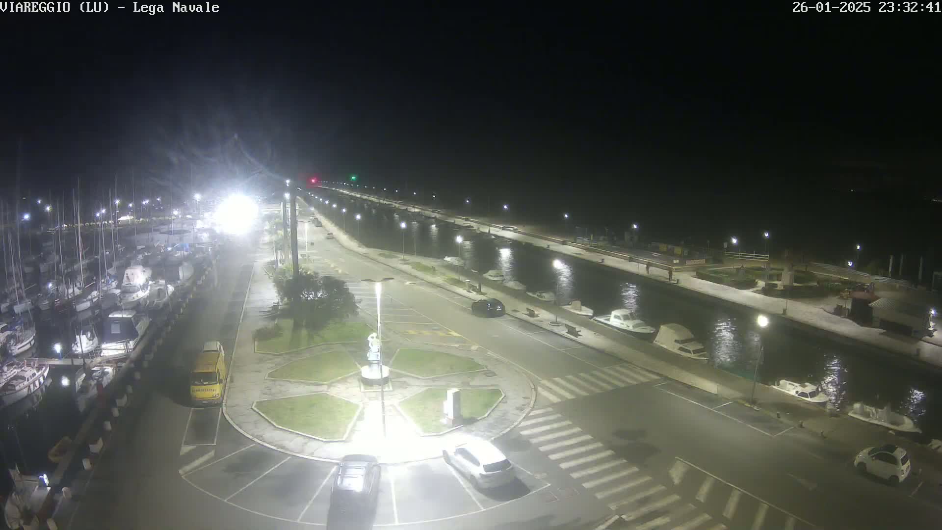 A nighttime view of a marina with numerous boats docked, a roadway lined with parked cars, and a small roundabout in the foreground, under clear skies.