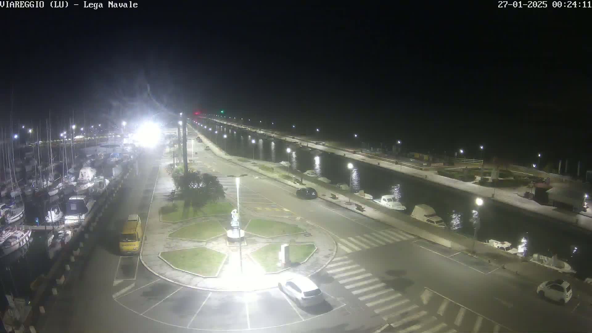 A nighttime view of a marina with numerous boats docked, a roadway with cars, and a small roundabout, all brightly lit under a dark, clear sky.