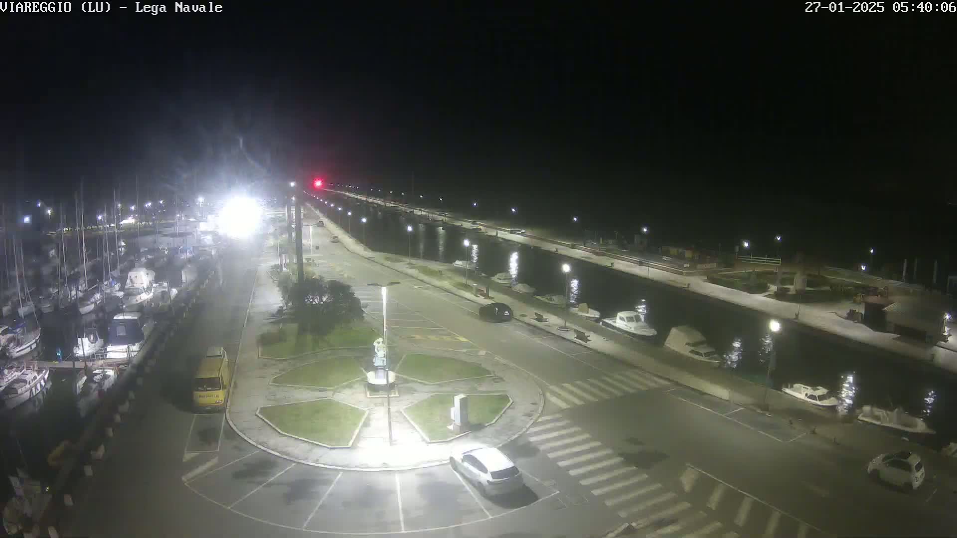 A nighttime, illuminated view of a marina with numerous boats docked, a roadway with cars, and a small, central plaza.