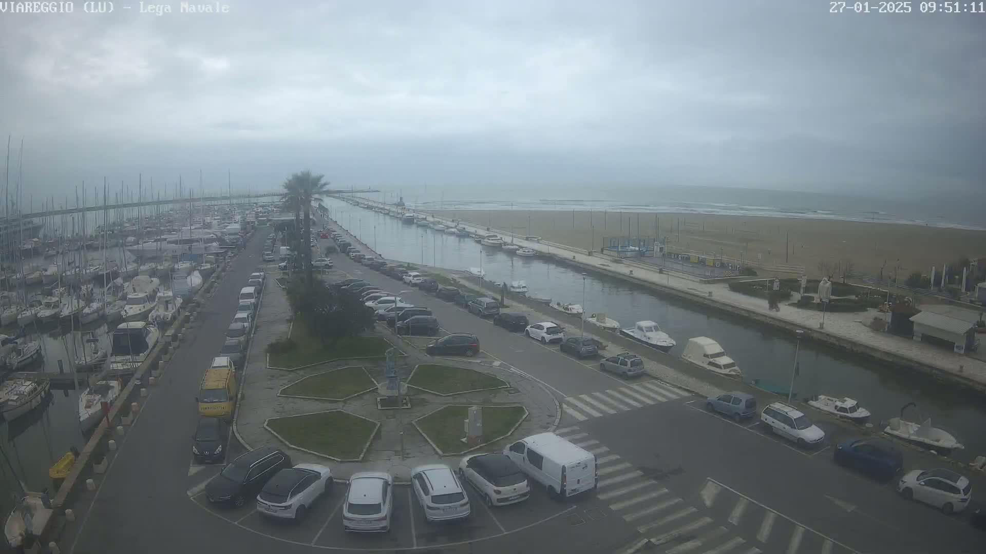 An overcast day shows a marina filled with boats, a canal lined with parked cars, and a sandy beach beyond.