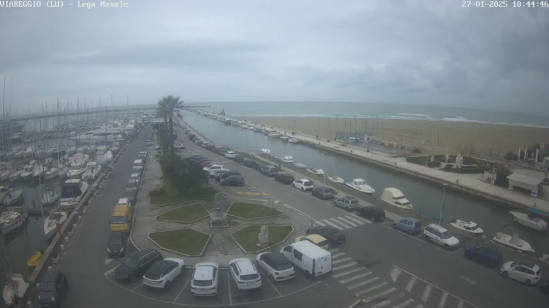 Here is a description of the image: An overcast day shows a marina filled with boats, a canal, a sandy beach, and a parking lot full of cars.