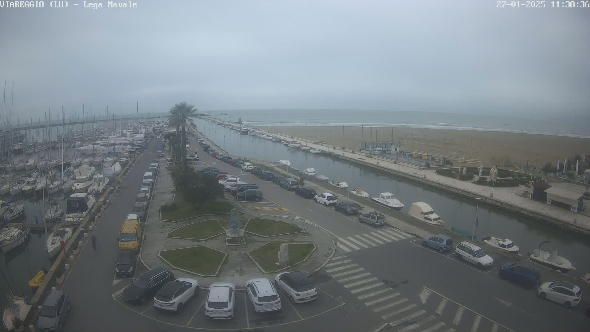 An overcast day shows a marina filled with boats, a canal lined with parked cars, and a sandy beach beyond.