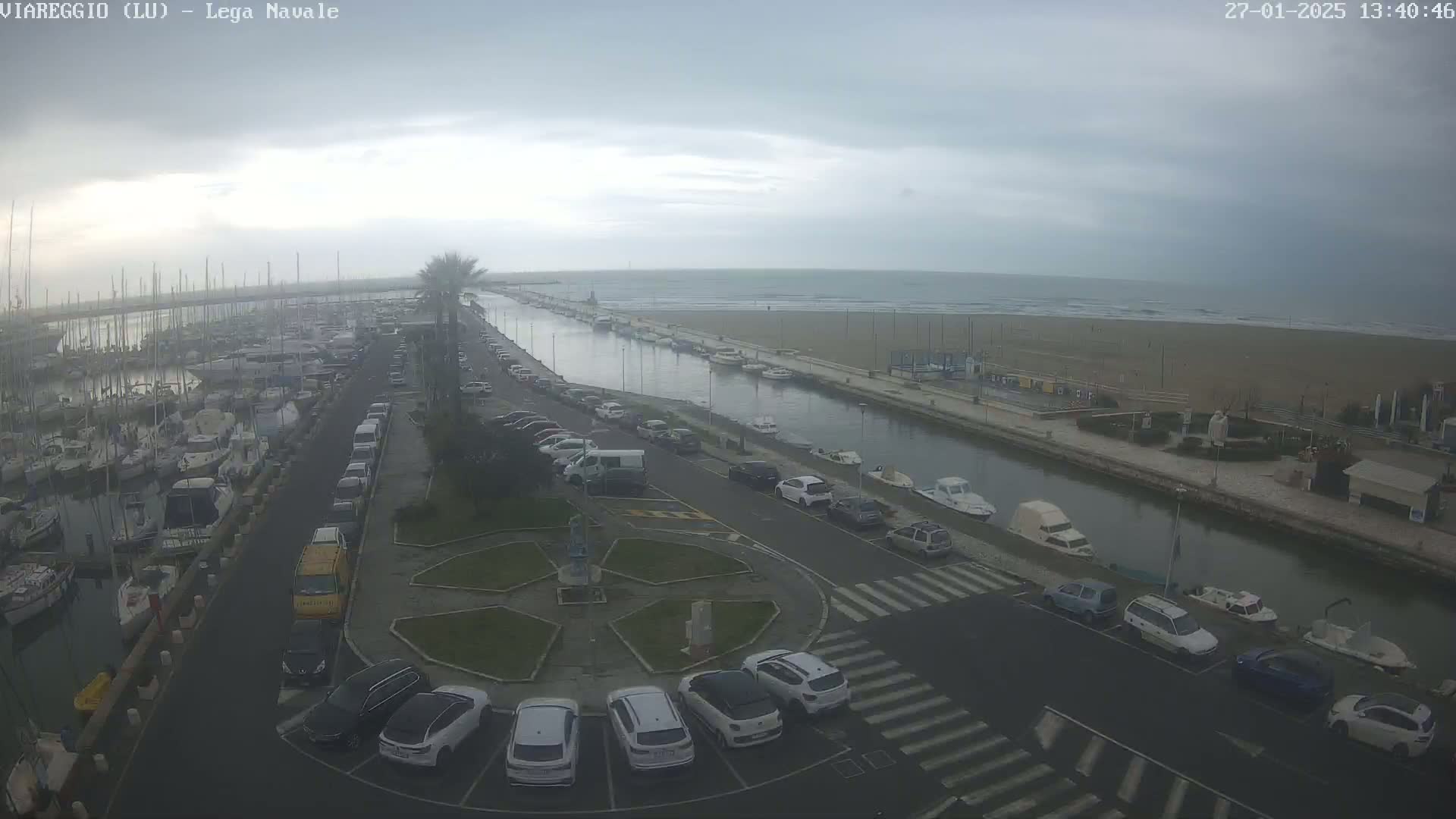 An overcast day reveals a marina filled with boats, a canal leading to a sandy beach and ocean, and a parking lot with cars surrounding a small, landscaped roundabout.