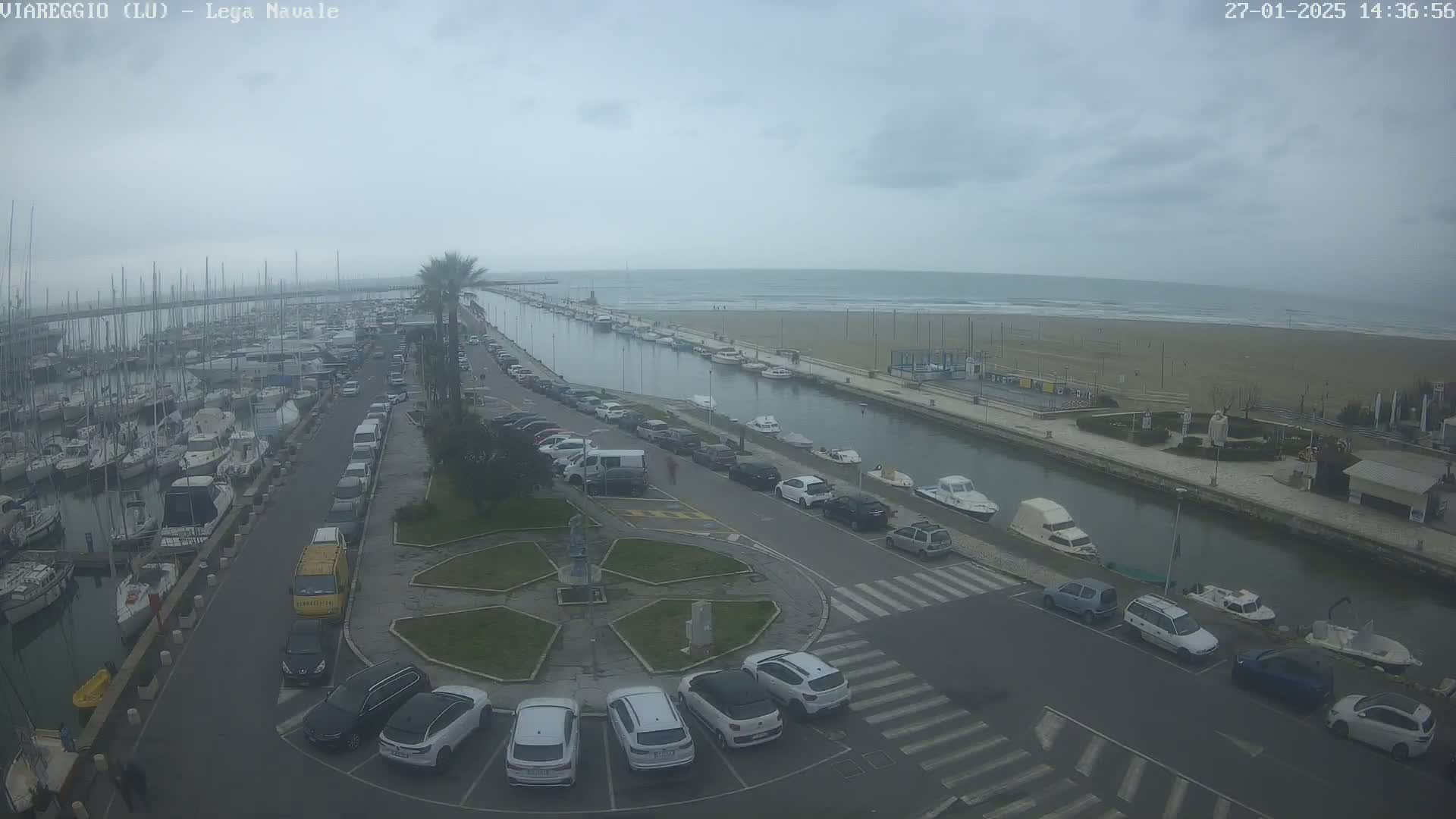 A marina filled with boats is next to a canal and a sandy beach under an overcast sky.