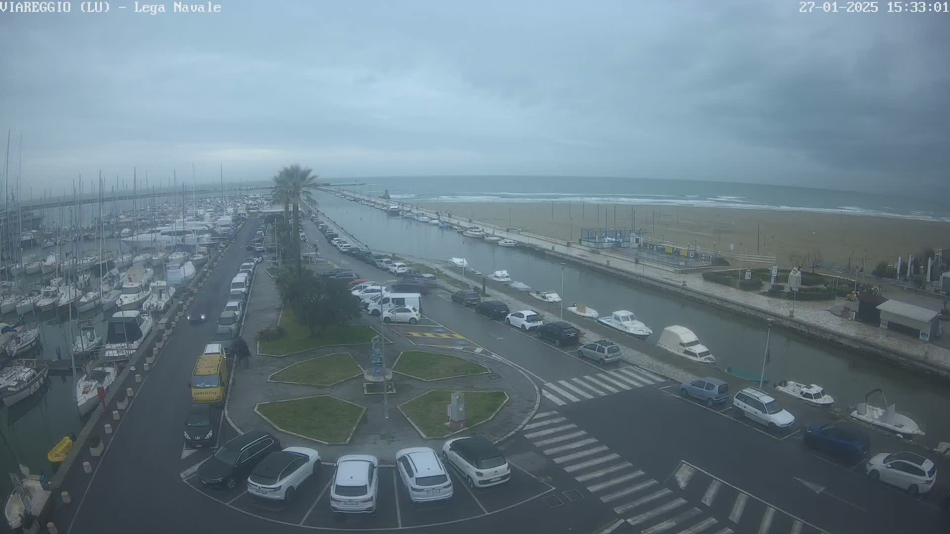 A harbor filled with boats, adjacent to a parking lot and canal leading to a beach with overcast skies and a wavy ocean.