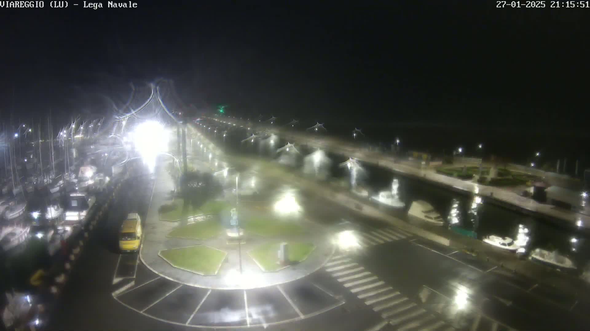 A nighttime, rain-soaked view of a harbor with boats docked, a roadway with vehicles, and illuminated walkways.