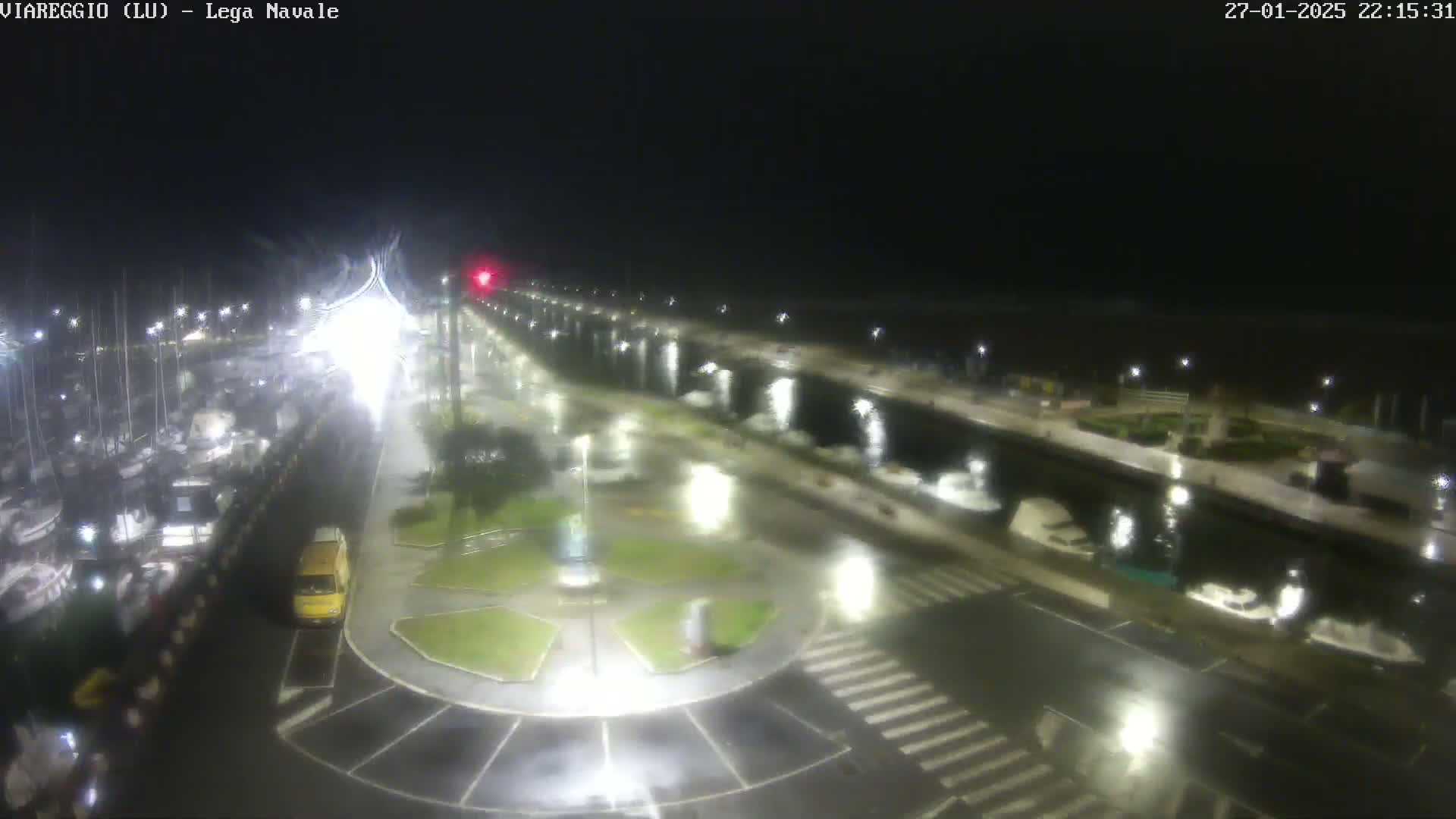 A wet, nighttime view of a marina with boats docked along a paved walkway lined with lights.