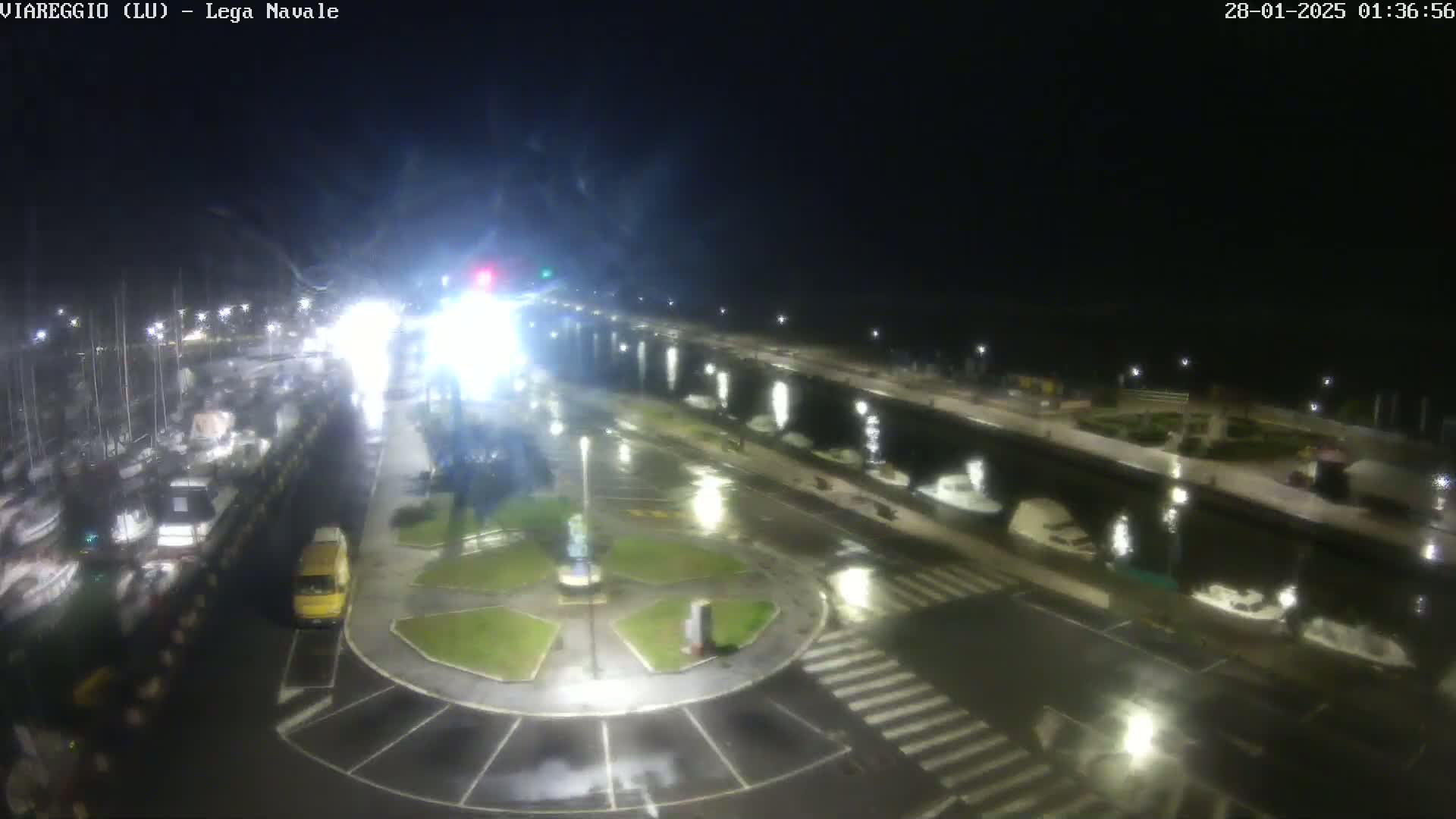 A wet, nighttime view of a harbor with boats docked alongside a roadway, featuring a small roundabout with a yellow van parked nearby.