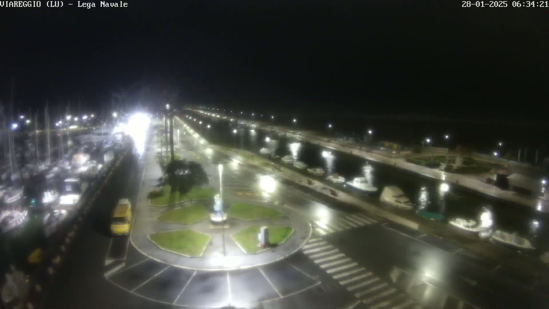 A nighttime, rain-slicked marina scene shows a harbor filled with boats and a roadway with a central roundabout, all illuminated by streetlights.