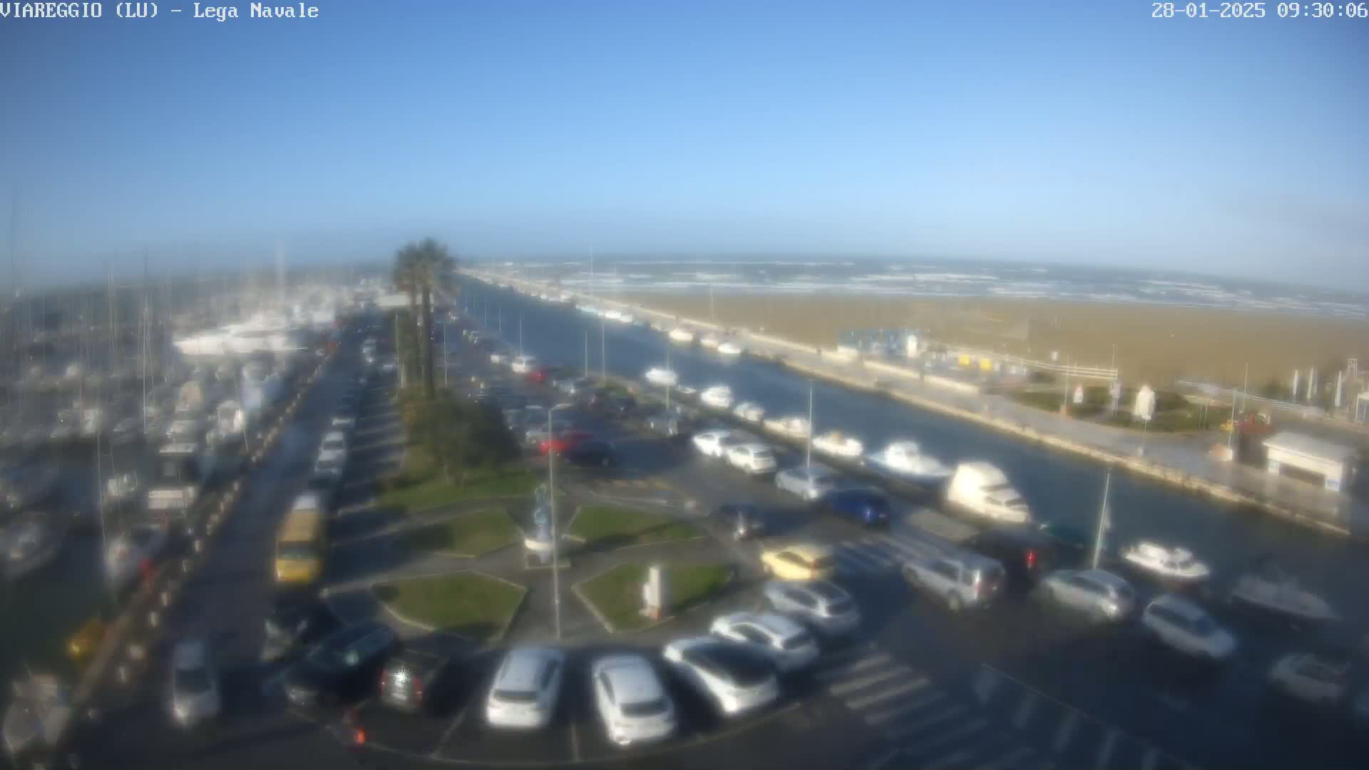 A marina filled with boats is adjacent to a canal and a beach with choppy waves under a clear blue sky.