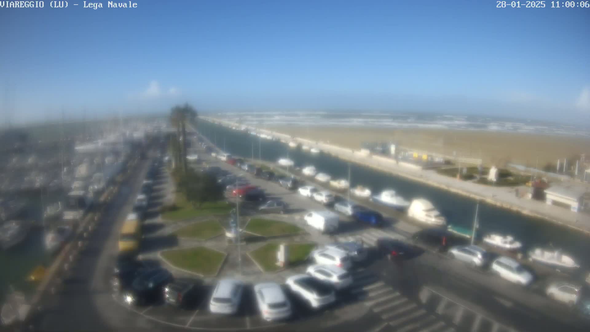 An aerial view shows a marina filled with boats, a parking lot with many cars, and a canal leading to a beach with choppy waves under a mostly clear sky.