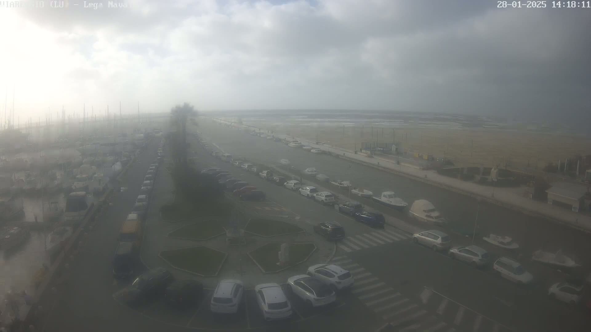 A hazy, overcast day reveals a harbor filled with boats, a parking lot with cars, and a beach in the distance.