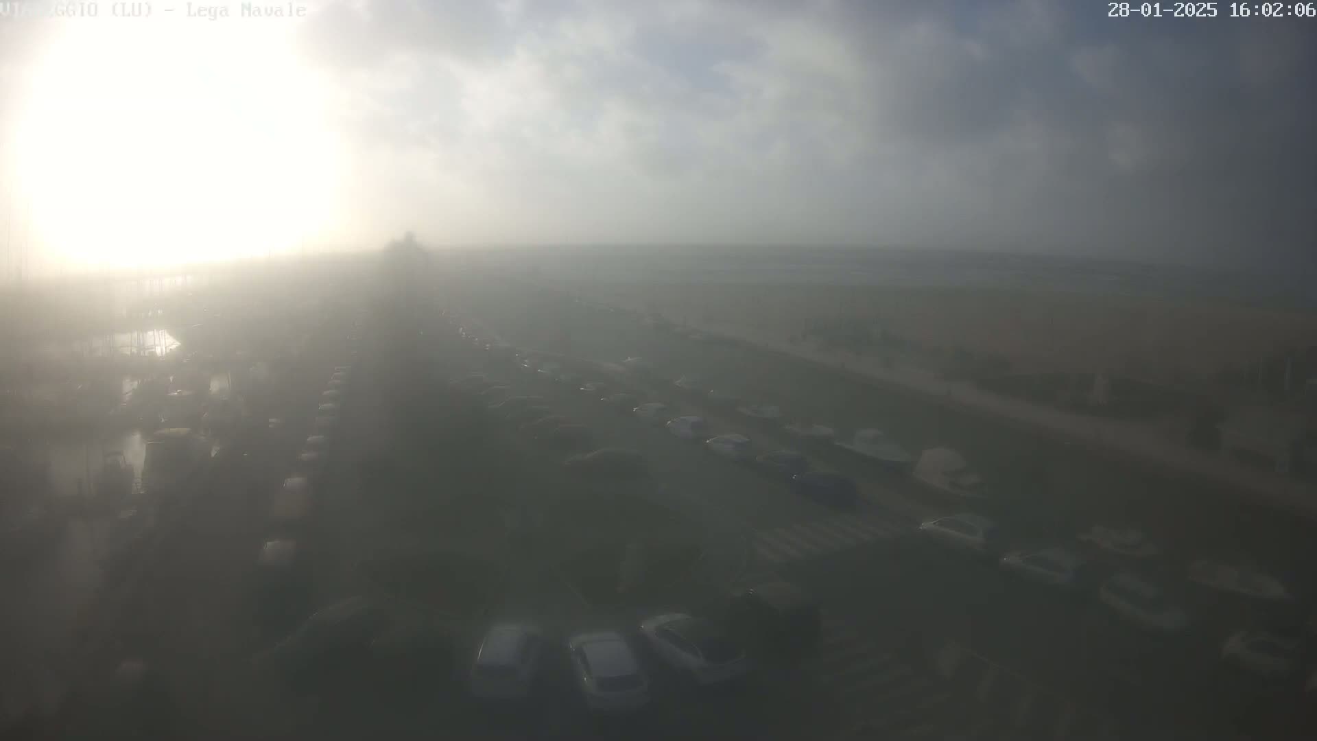 A hazy, sunlit scene shows a large parking lot and a roadway filled with numerous vehicles, extending towards a foggy waterfront.