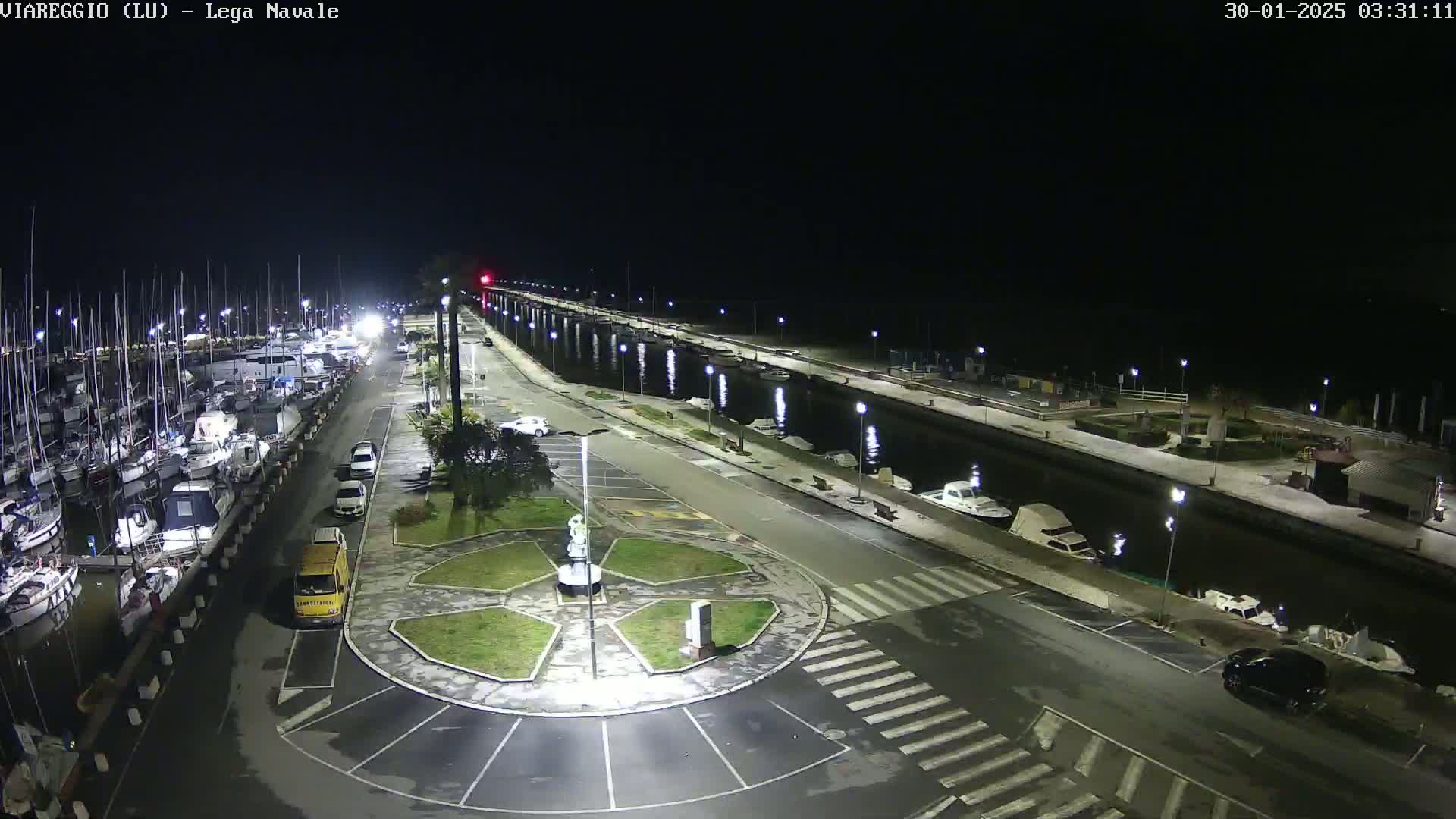 A nighttime aerial view shows a marina filled with boats, a roadway with parked cars and a central roundabout, all illuminated by streetlights.