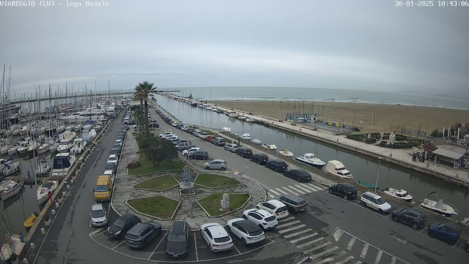 An overcast day shows a marina filled with sailboats and motorboats, a canal lined with parked cars, and a sandy beach beyond.