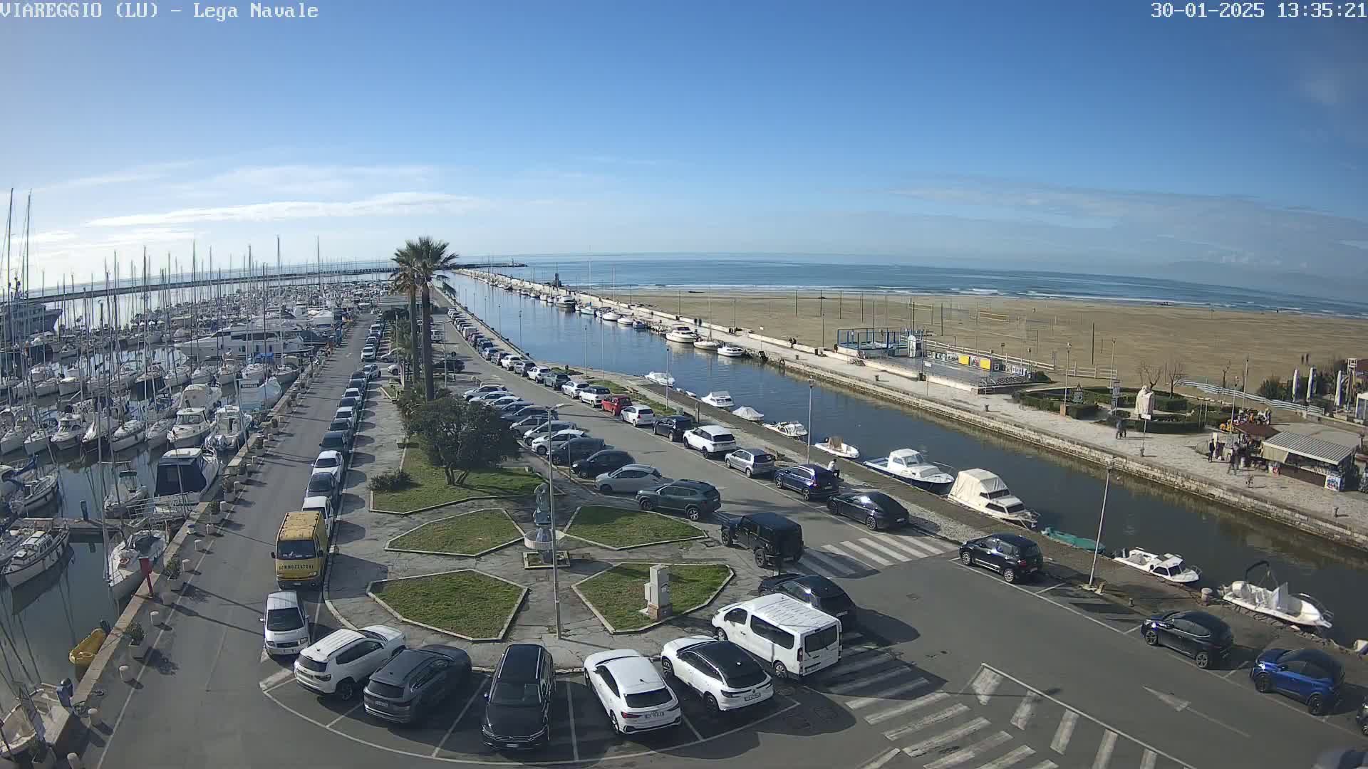 A marina filled with boats and surrounded by cars parked along a canal that leads to a sandy beach under a clear blue sky.