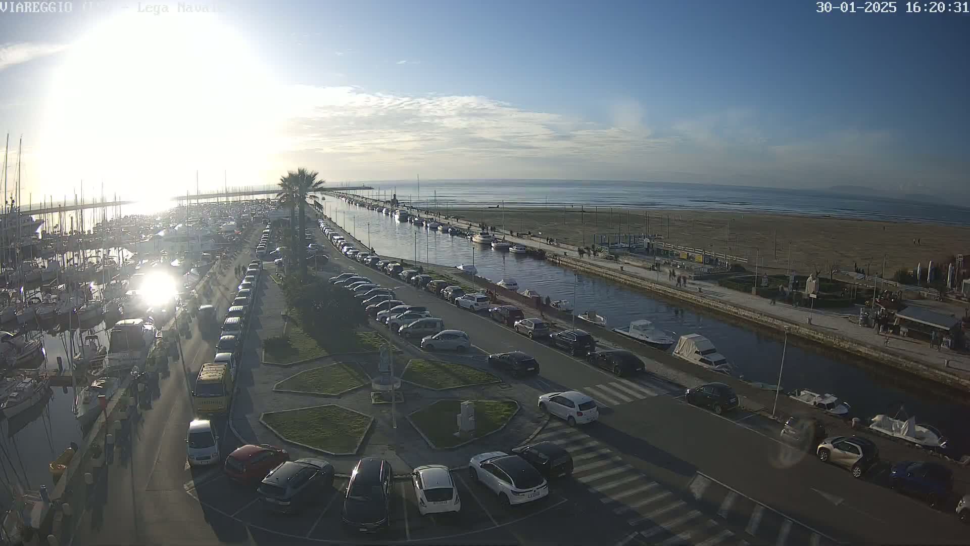 A marina filled with boats and surrounded by parked cars is next to a canal that leads to a sandy beach and calm ocean under a mostly sunny sky.