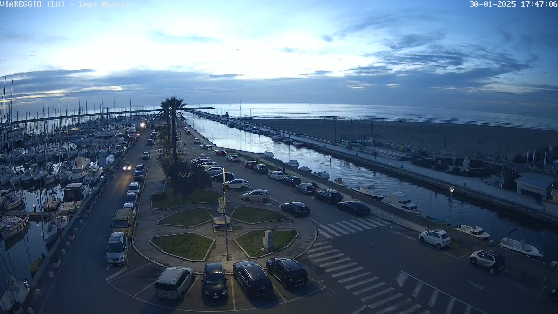 A marina filled with boats is adjacent to a canal that leads to an ocean beach under a twilight sky.