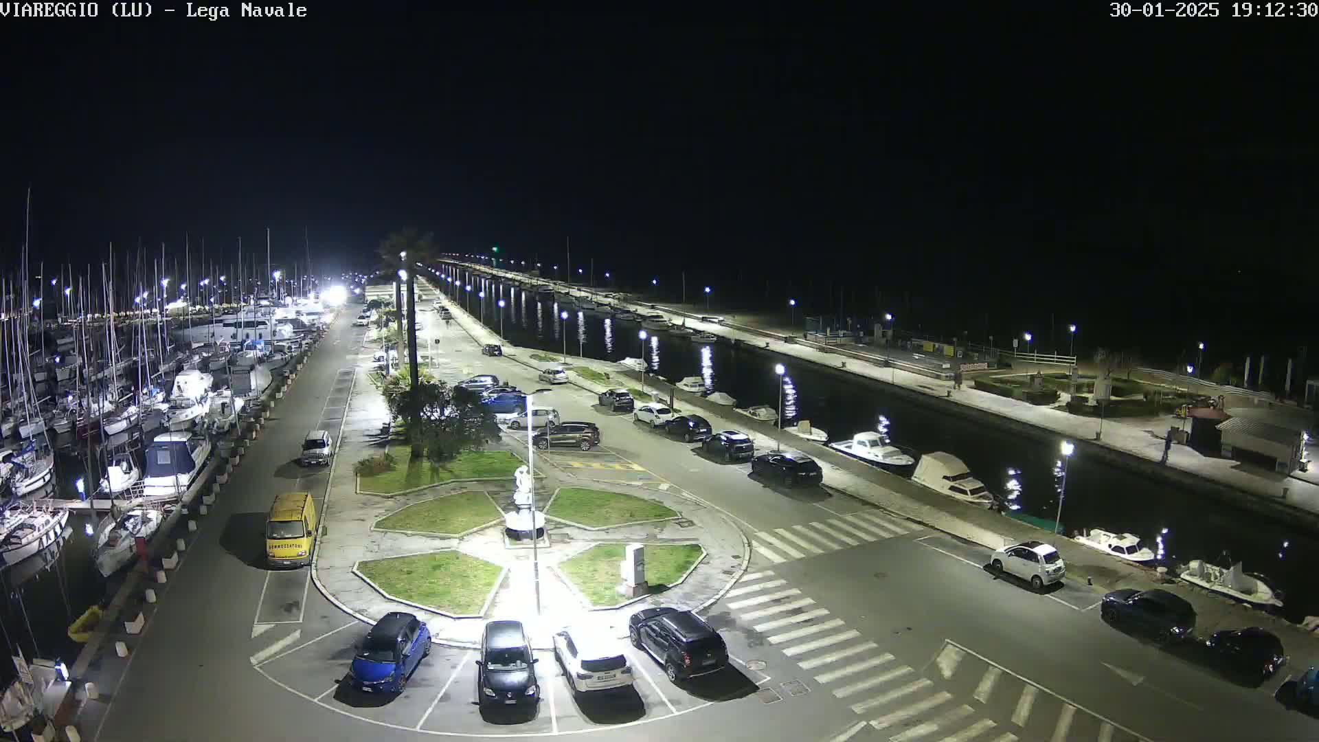 A nighttime aerial view of a marina with boats docked, cars parked in a lot surrounding a central fountain, and a canal running alongside a paved walkway.