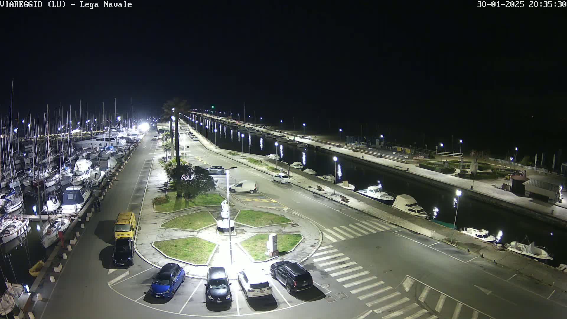A nighttime aerial view of a marina with numerous boats docked, cars parked in a lot, and a canal lined with lights.