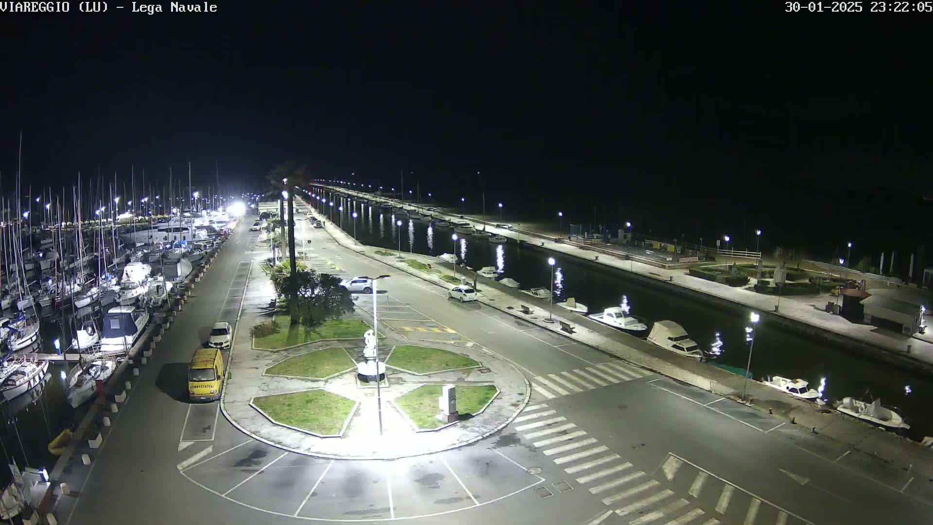 A nighttime aerial view of a marina with numerous boats docked, a canal, and a circular plaza with a statue in the center, illuminated by streetlights.