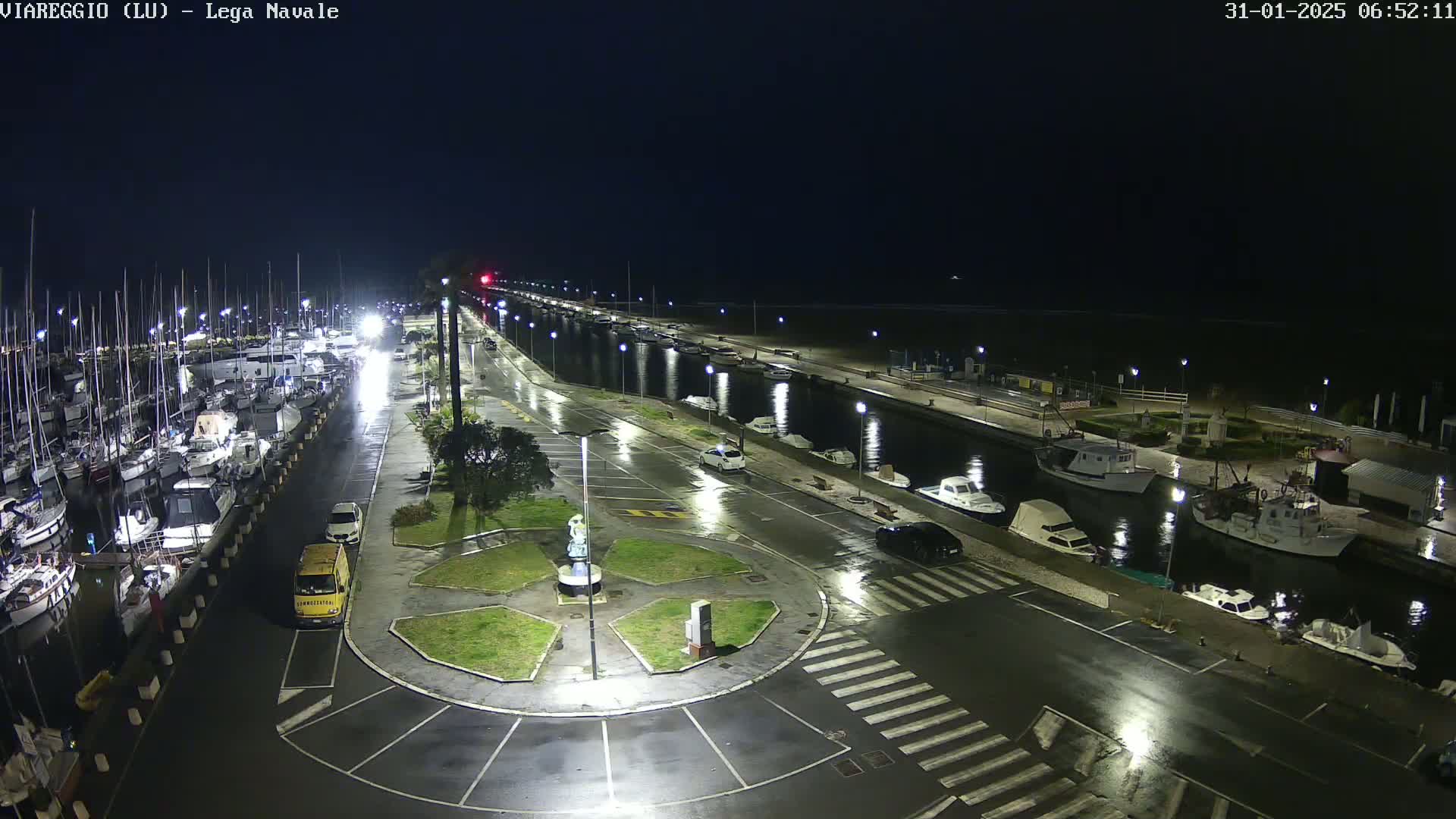 A nighttime view of a marina with many boats docked, wet roads, and a few cars parked, under rainy conditions.