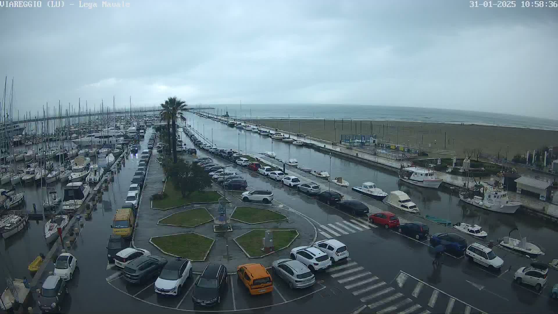 A marina filled with boats is partially submerged in floodwater under a cloudy sky, with cars parked along a canal next to a beach.