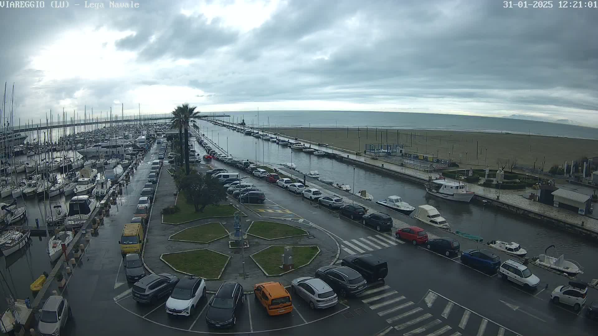 A wet, overcast day reveals a marina filled with boats, a canal lined with parked cars, and a sandy beach under a cloudy sky.