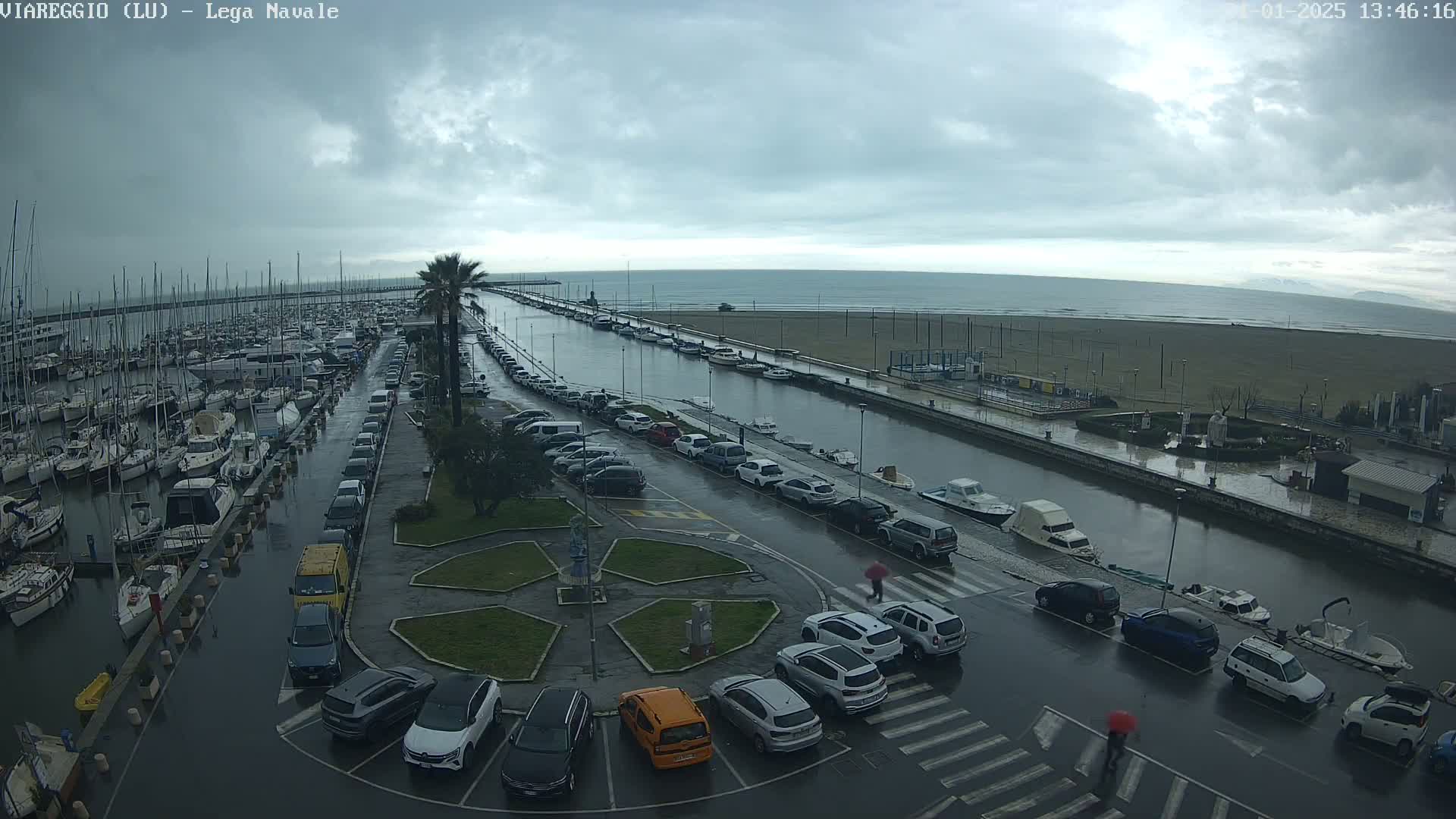 A wet marina filled with boats is adjacent to a wet parking lot and a canal leading to a beach under a cloudy sky.