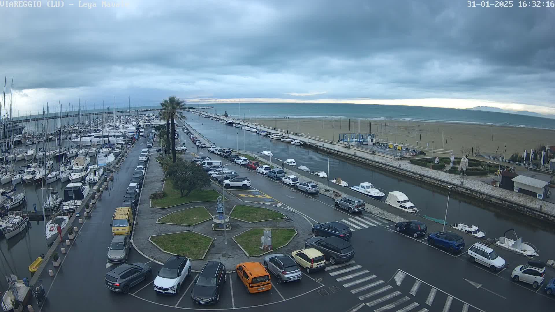 A harbor filled with boats is next to a sandy beach under an overcast sky, with a canal separating the harbor from the beach and parking lots lining the canal.