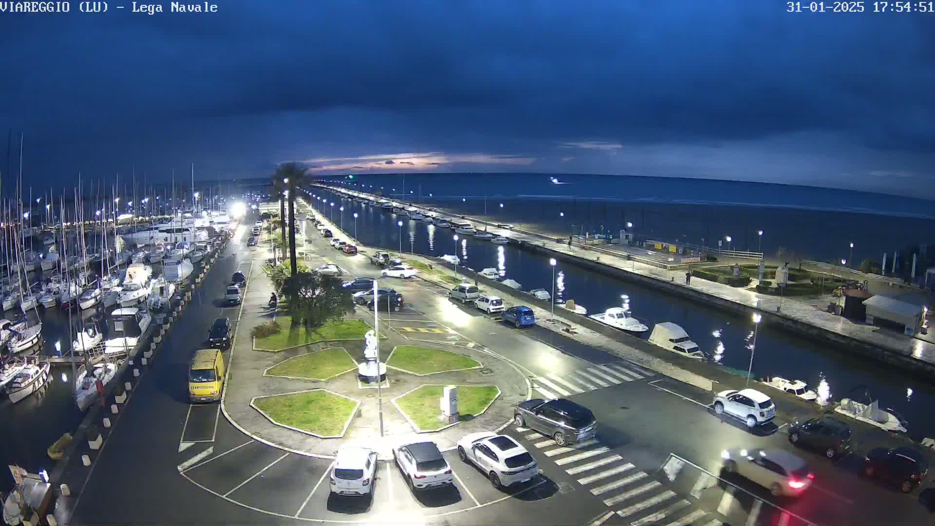 A marina at twilight under a partly cloudy sky, with cars parked along a waterfront road and boats docked in the harbor.