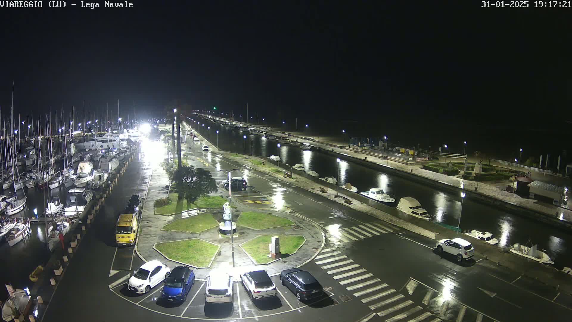 A nighttime view of a marina with numerous boats docked, a wet paved area with cars parked, and a canal with additional boats.