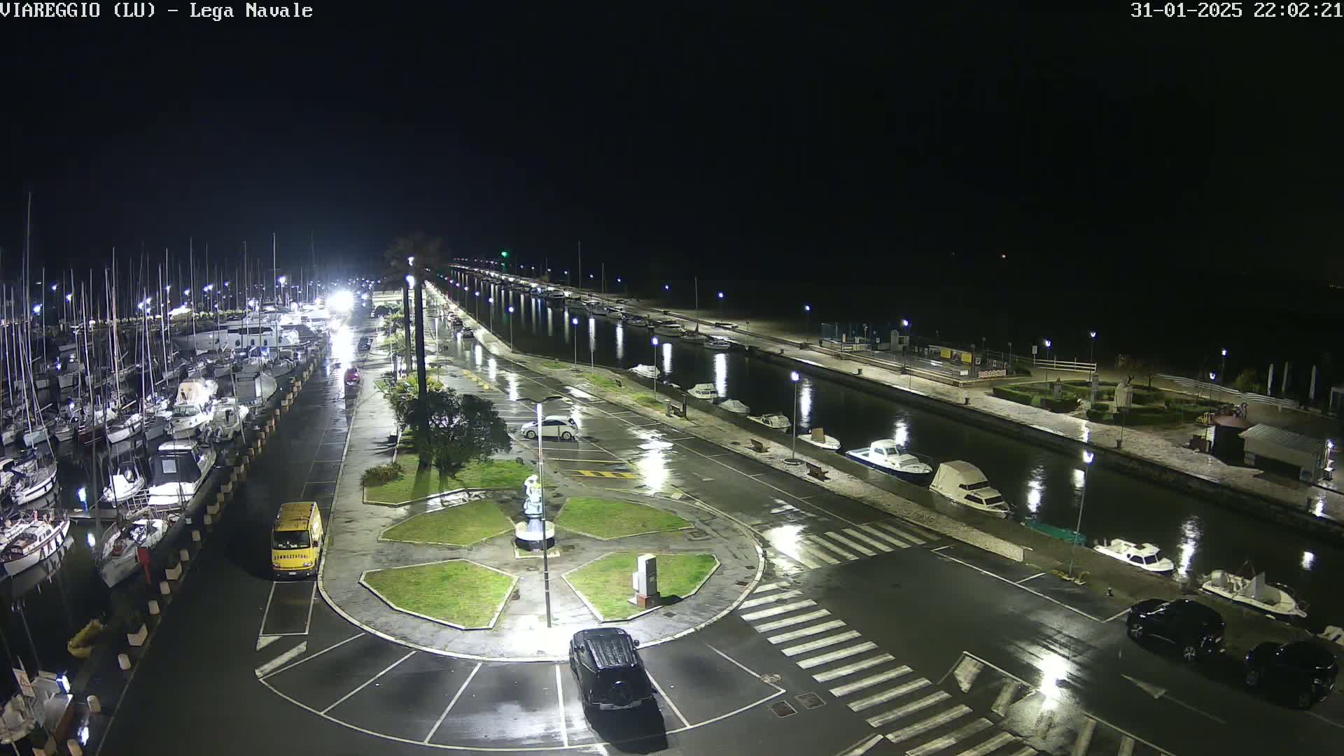 A nighttime view of a marina with numerous boats docked, wet roads, and several cars parked, under rainy conditions.