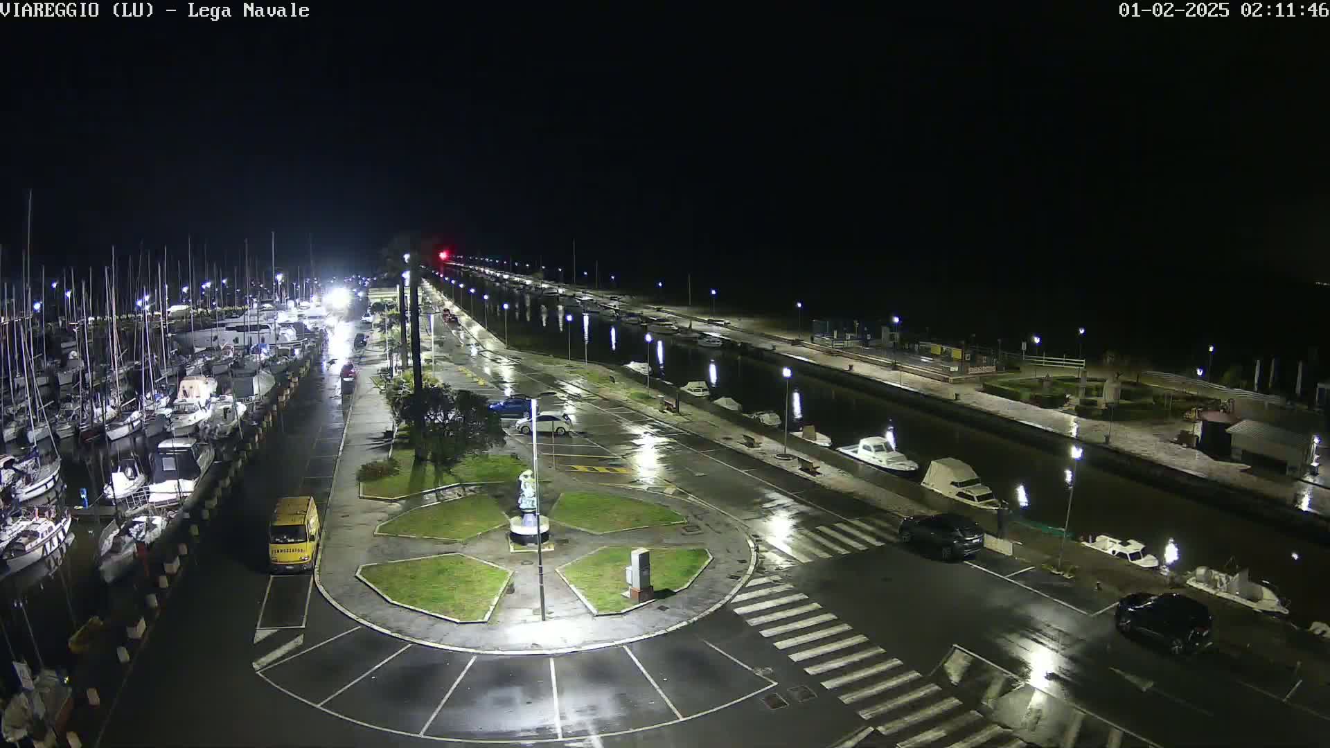 A nighttime, rain-slicked marina and roadway scene shows numerous boats docked, cars parked, and a small roundabout with a statue in the center.