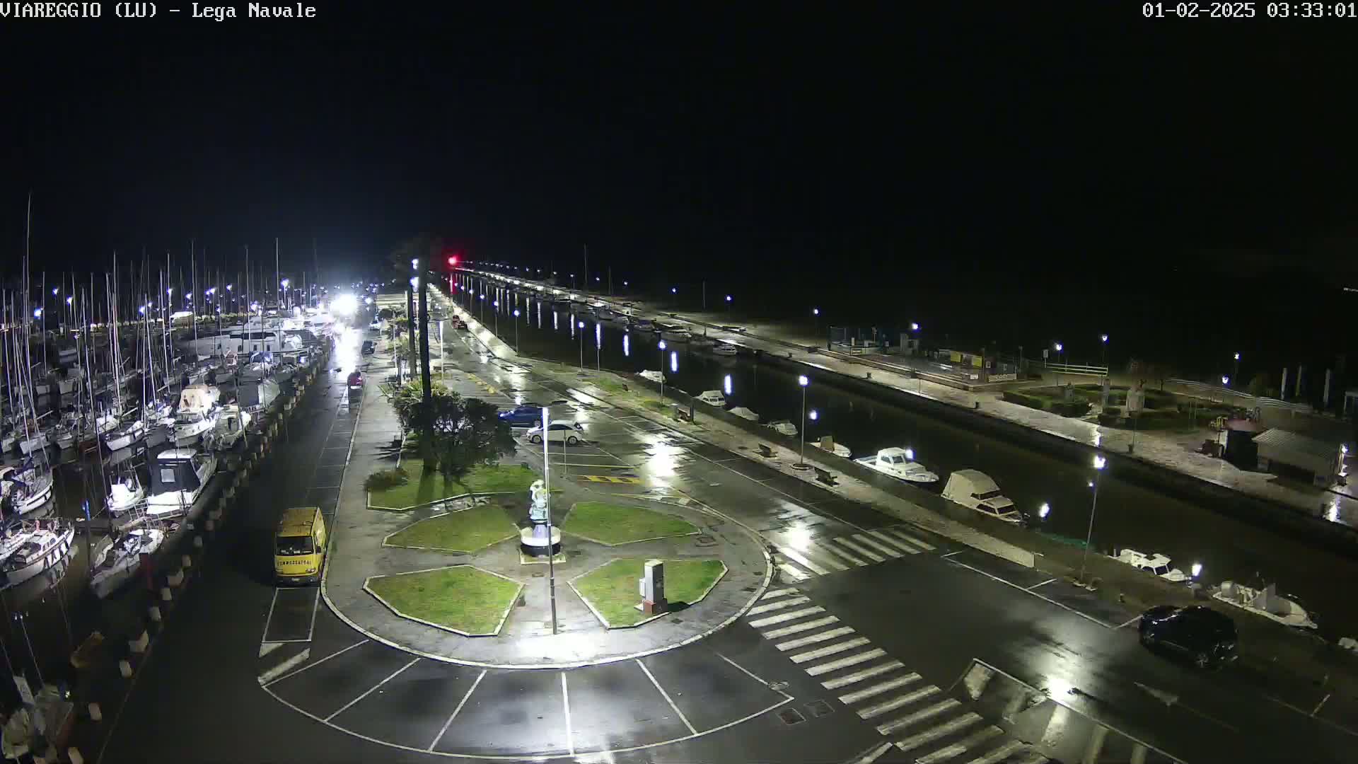 A nighttime, rain-slicked marina with numerous boats docked, a few cars parked, and a small roundabout in the foreground.