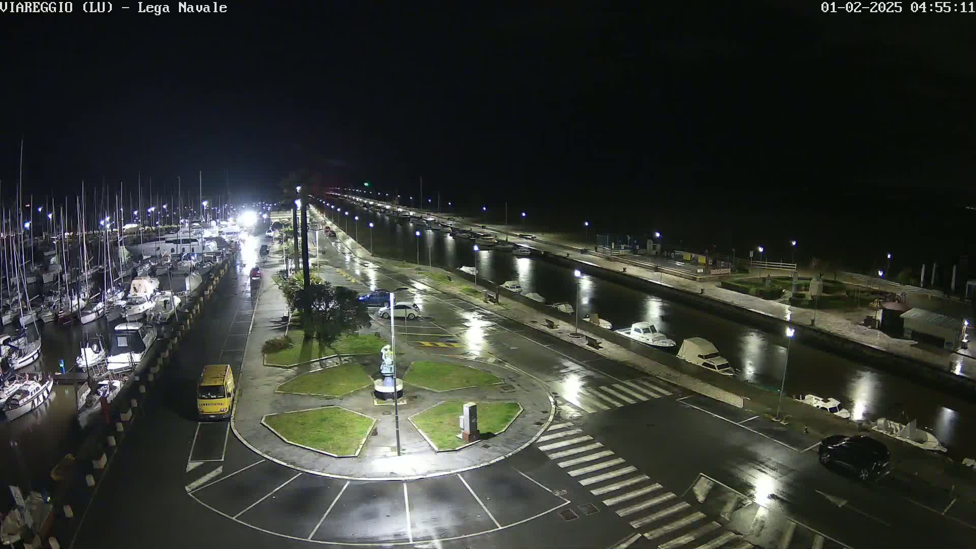 A nighttime, wet marina with boats docked along a canal, cars parked in a lot, and a small roundabout in the center of the roadway.
