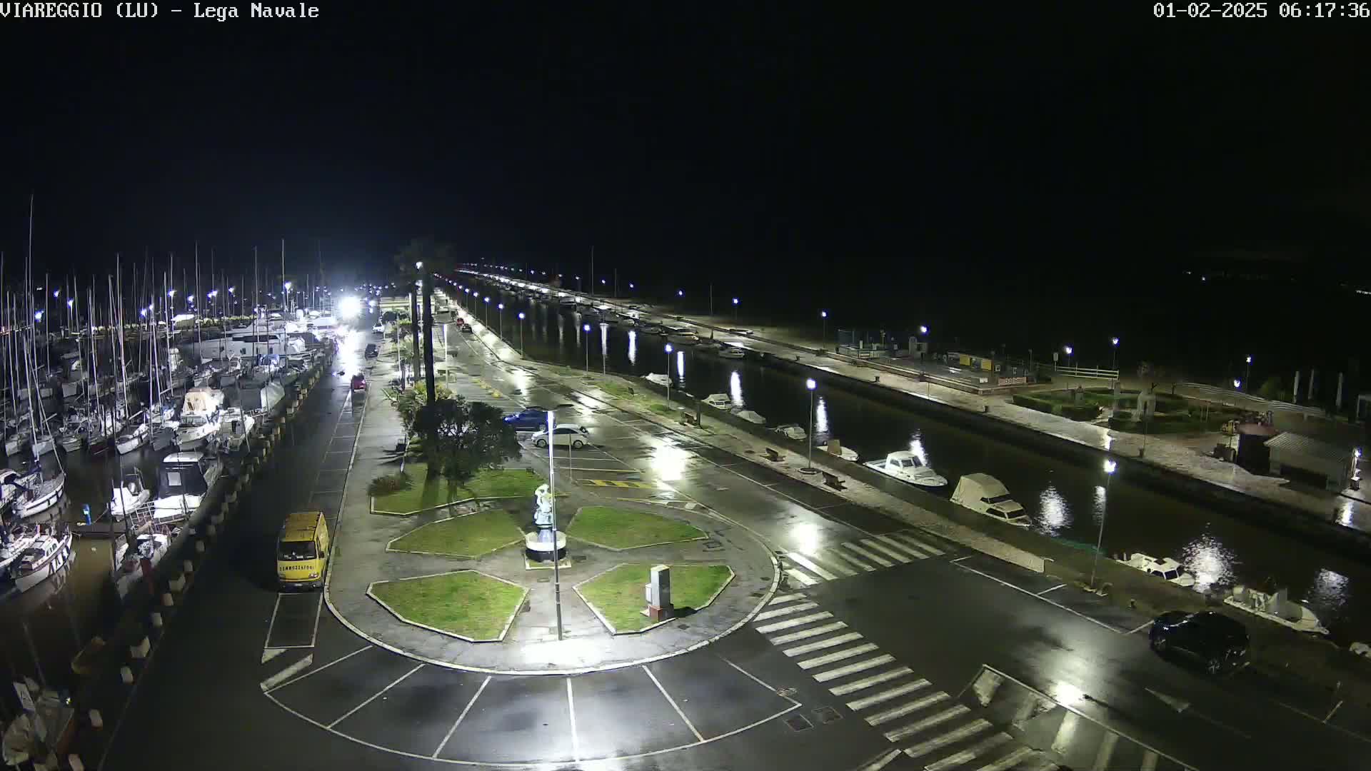 A nighttime, wet marina scene shows numerous boats docked, a canal with additional boats, and a wet roadway with several cars parked.