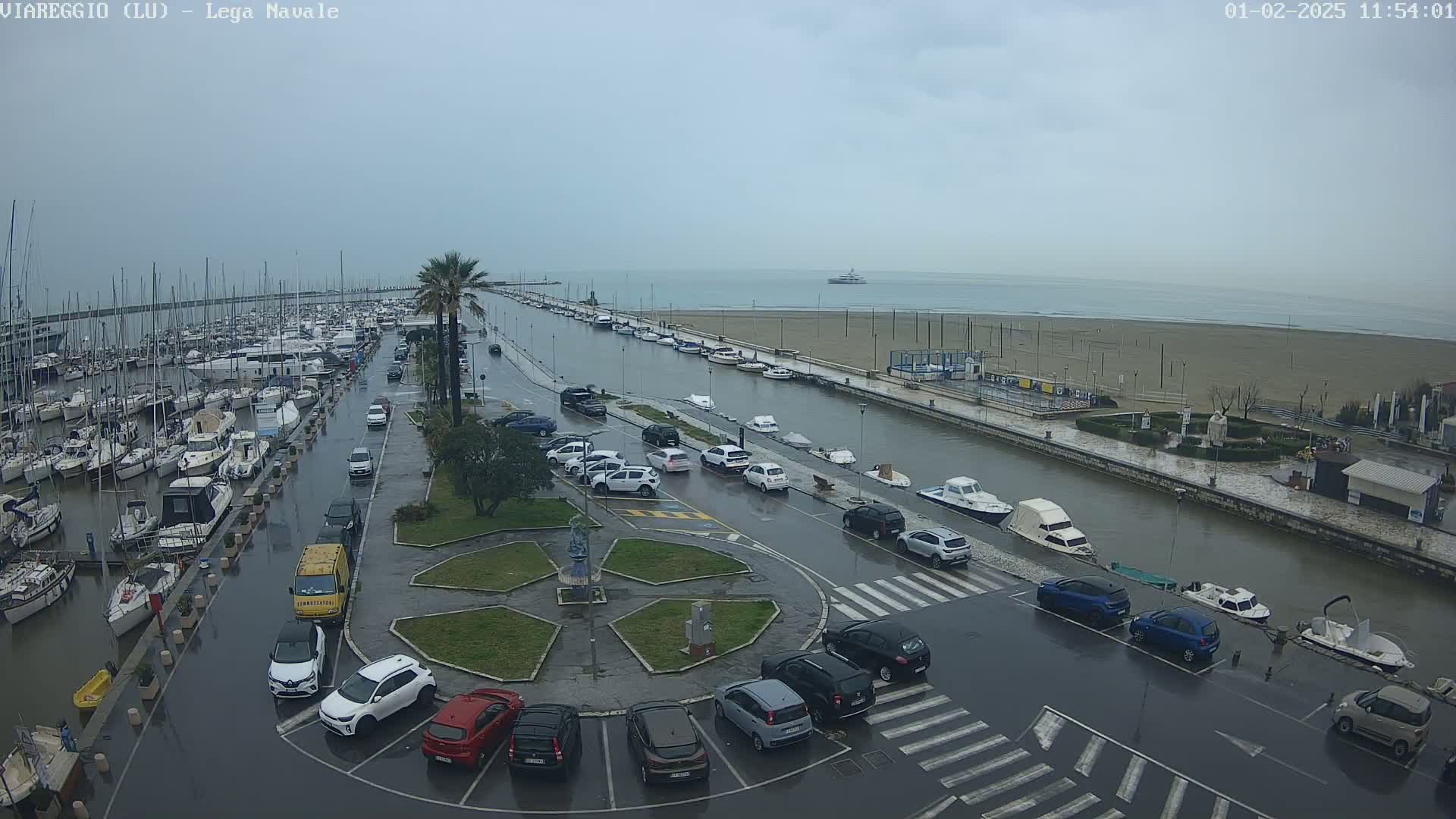 A marina with many boats is partially flooded, with cars parked along a canal-like waterway under an overcast sky.
