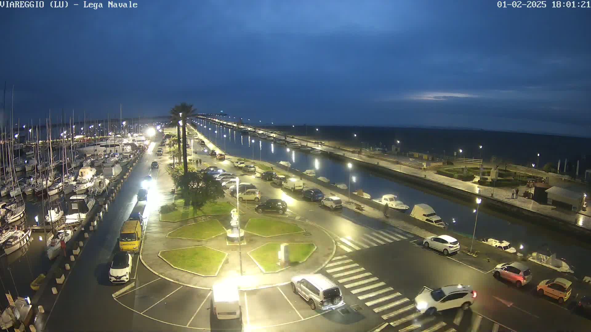 A marina at night under a twilight sky, with cars parked along a canal next to a waterfront promenade.