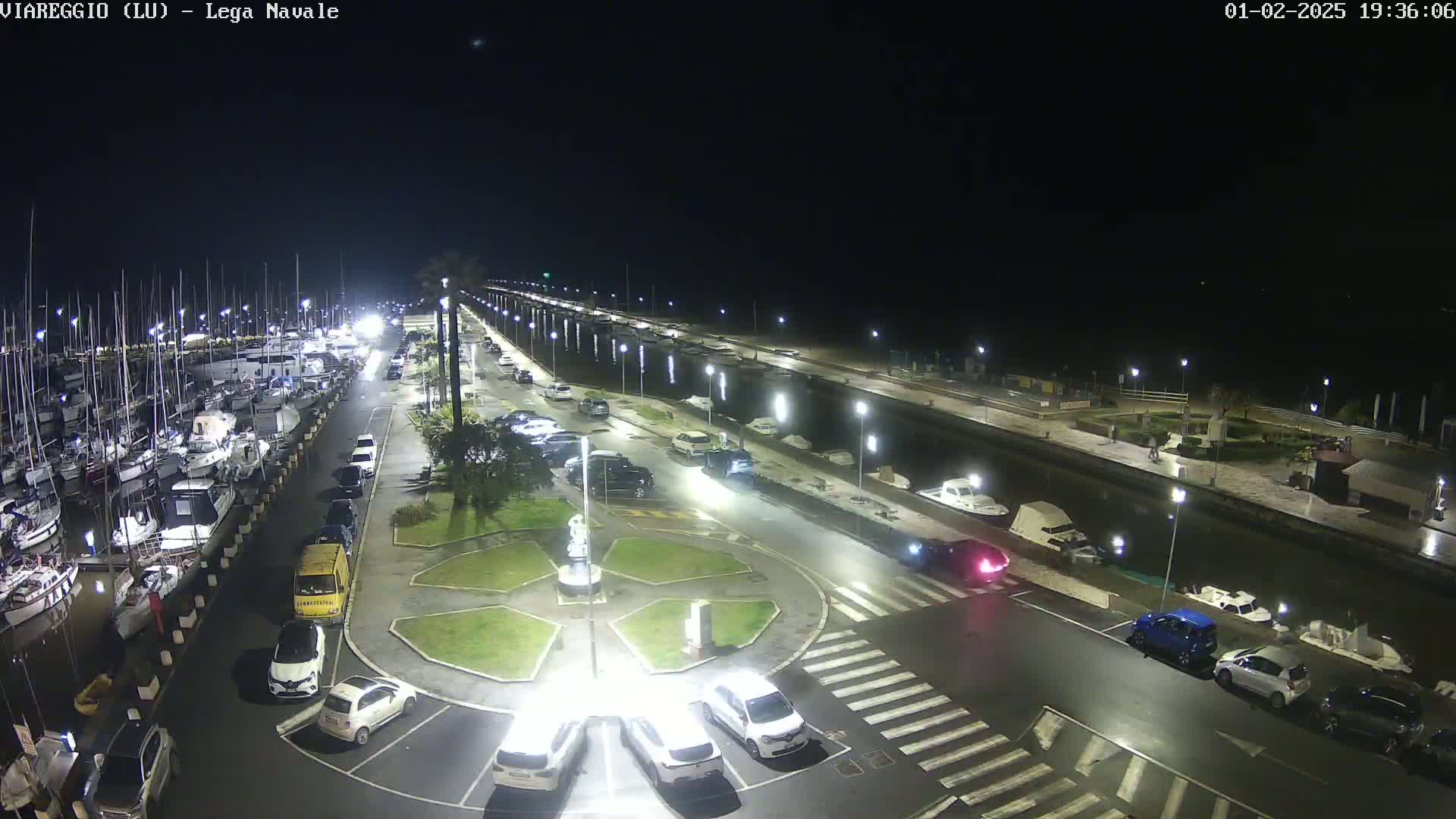 A nighttime aerial view of a marina with boats docked, cars parked in a lot, and a roadway leading to a roundabout.