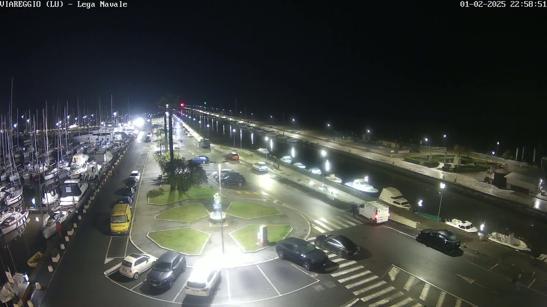A nighttime aerial view of a marina with numerous boats docked, a roadway lined with parked cars, and a central roundabout.
