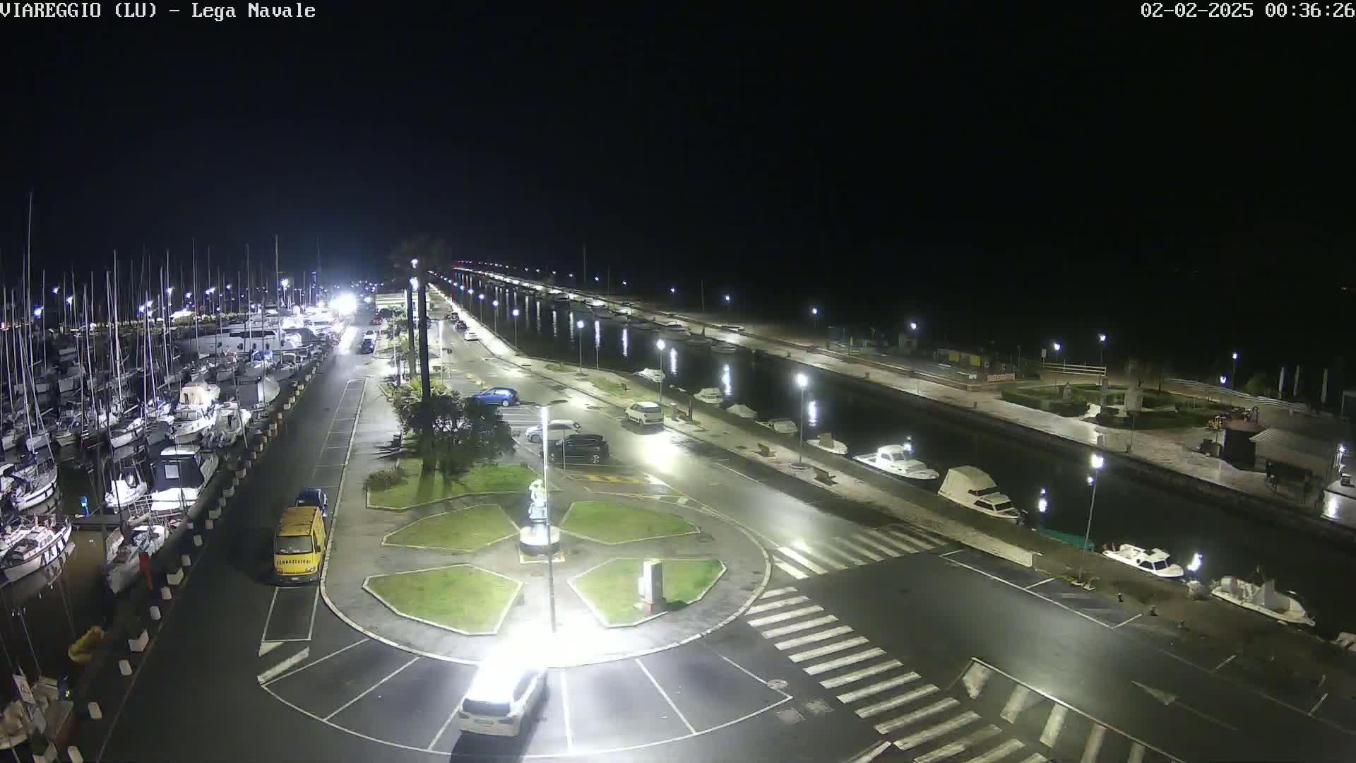 A nighttime, aerial view of a marina filled with boats, cars parked in a lot nearby, and a roundabout in the foreground.