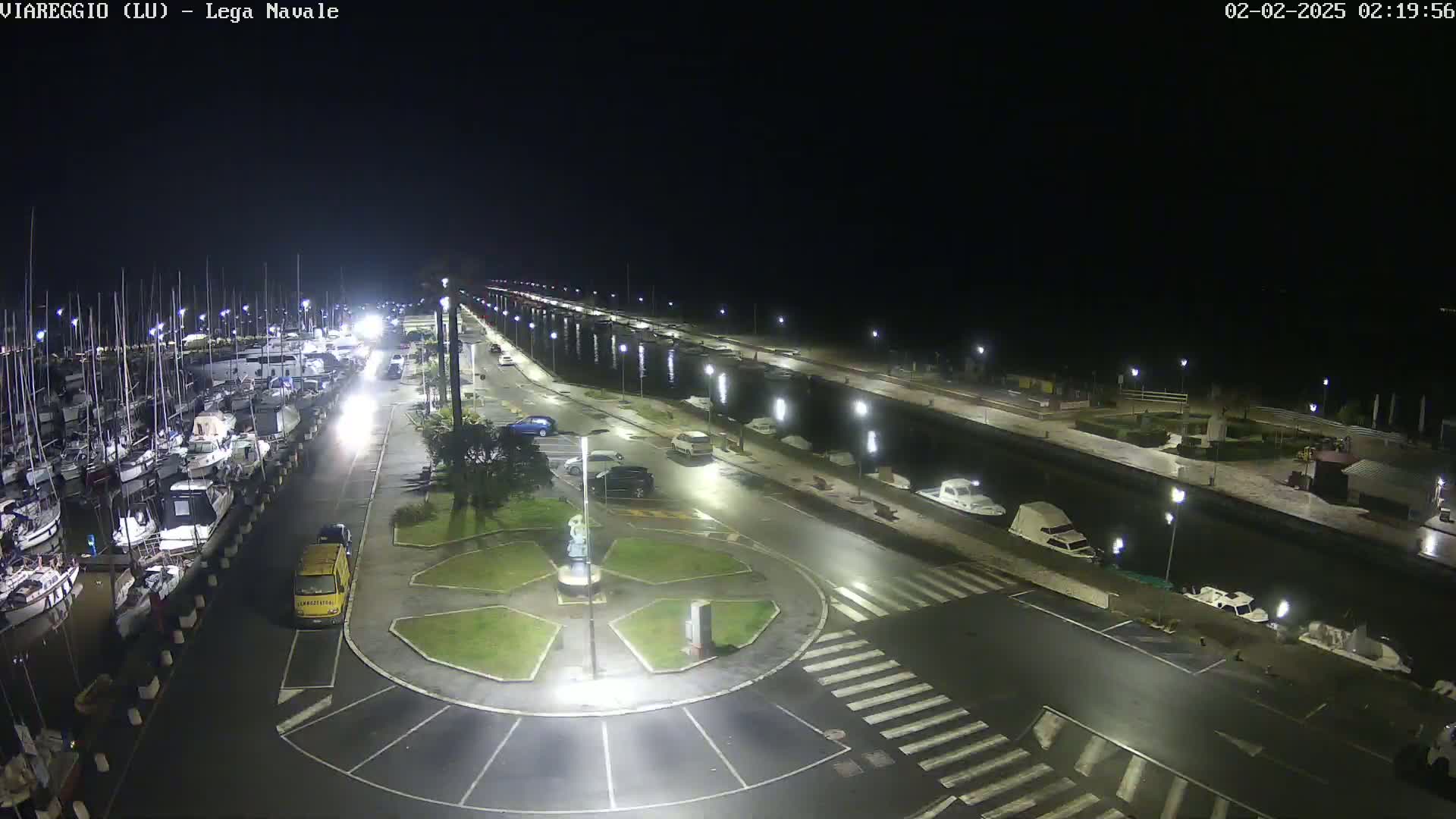 A nighttime view of a marina with many boats docked, a roadway with parked cars, and a central roundabout, all illuminated by artificial lights.