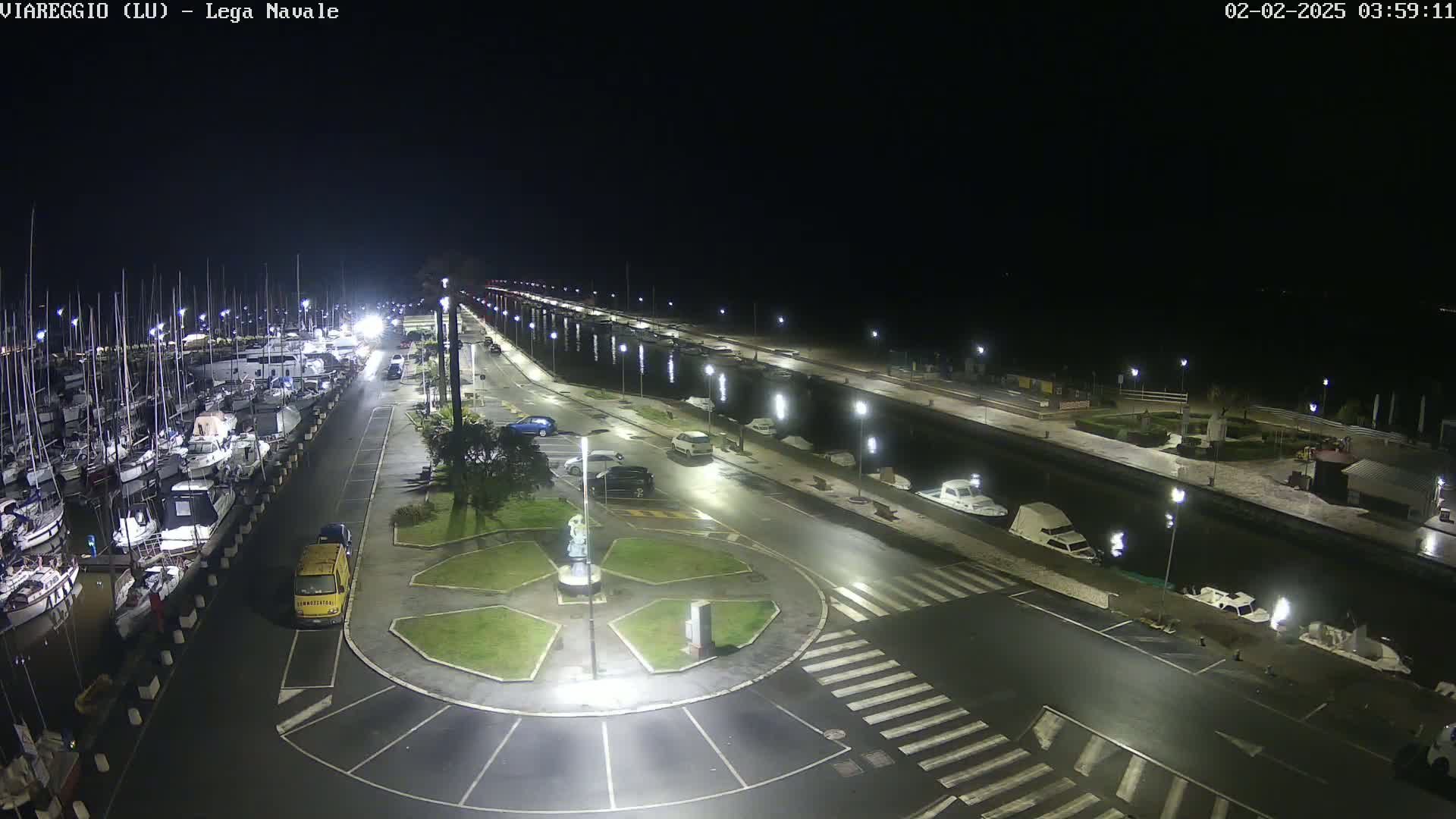 A nighttime, aerial view of a marina with many boats docked, a roadway with parked cars, and a central roundabout, all illuminated by streetlights.