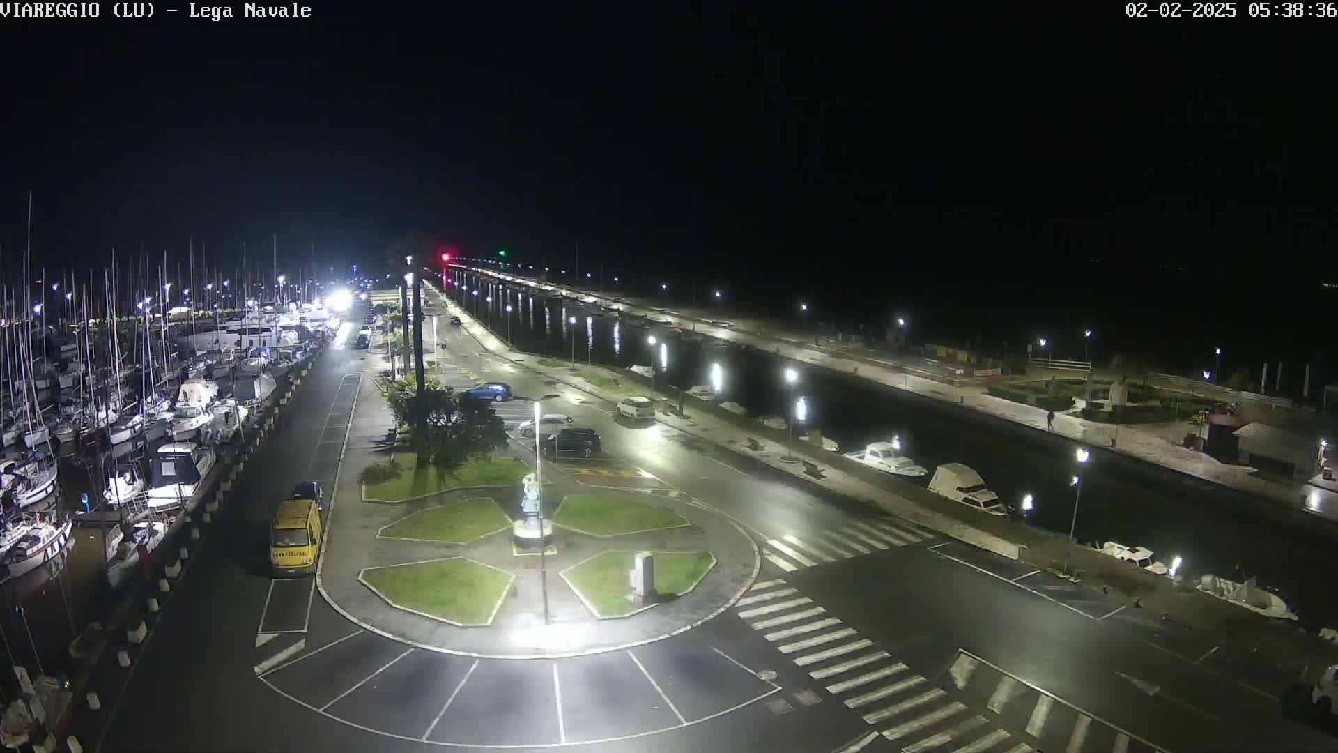 A nighttime aerial view of a marina with numerous boats docked, a roadway with parked cars, and a central roundabout, all brightly lit.