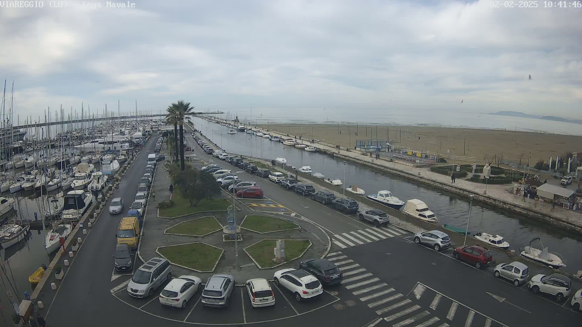 An overcast day reveals a marina filled with boats, a canal lined with parked cars and small boats, and a sandy beach beyond.